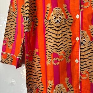 May include: Orange and pink striped pajama top with a repeating tiger print. The tigers are tan with black stripes. The top has white trim and white buttons.