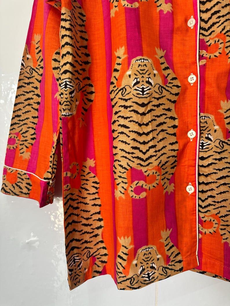 May include: Orange and pink striped pajama top with a repeating tiger print. The tigers are tan with black stripes. The top has white trim and white buttons.