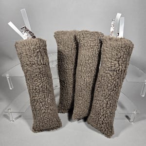 May include: Four brown, cylindrical, plush fabric items with white ribbon and patterned accents. The items are arranged on a clear, tiered display stand against a gray background. The plush fabric has a textured, fuzzy appearance.