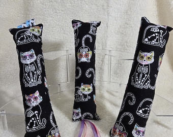 May include: Three black cat toys with a Day of the Dead theme. Each toy is a long, rectangular shape with a cat skeleton design and colorful floral accents. The toys are made of fabric and have a small ribbon attached.