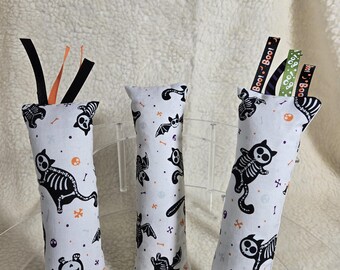 May include: Three Halloween-themed cat toys. Each cylindrical toy is white with a repeating pattern of black skeleton cats, bats, and orange accents. Black and orange ribbons are attached to the top of two toys, and the text "Boo!" is printed on the ribbons of the third.
