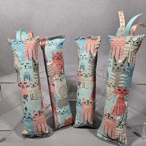May include: Four catnip-filled cat toys. Each rectangular toy is made of fabric with a repeating pattern of cat faces in pink, blue, gray, and white. The toys have a ribbon attached to the top with paw prints.