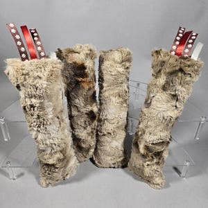 May include: Four faux fur Christmas stockings in varying shades of brown and gray. Each stocking has a decorative ribbon with paw prints and a red ribbon. The stockings are displayed on a clear, tiered stand.