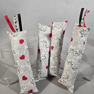 May include: Four rectangular fabric cat toys featuring a light gray background with a repeating pattern of red hearts and the text Crazy Cat Lady in black script. Each toy is topped with three short ribbons in red, black, and white paw print patterns.