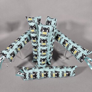 May include: Cat toys in a light blue fabric with a repeating pattern of black cat faces and the word "PEOPLE" in yellow. Each toy is a cylindrical shape with a ribbon attached. The toys are arranged on a clear stand.