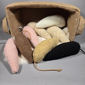May include: A collection of plush, ear-shaped toys in various colors, including pink, brown, yellow, and black, spilling out of a woven basket. Each toy has a leather string attached. The basket has a brown interior.