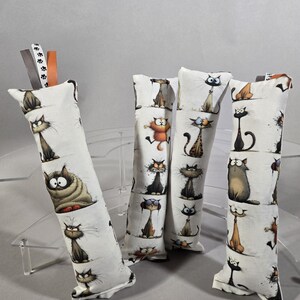 May include: Four rectangular fabric cat toys featuring a white background with a repeating pattern of various cartoon cats in different colors and expressions. Each toy has small colorful ribbon tags attached to the top edge for added texture.