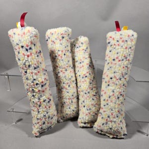 May include: Four rectangular, plush, off-white fabric tubes featuring a multi-colored confetti-like pattern of small red, blue, and yellow specks. Each tube is topped with a small, thin red ribbon loop extending from the top edge.