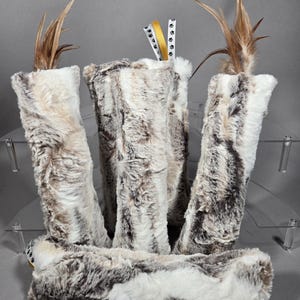 May include: A collection of plush, faux fur pet toys in shades of white, gray, and brown. Each toy is cylindrical, with some featuring a feather accent and ribbon with paw print details. The toys are arranged on a clear display.
