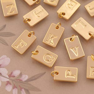 May include: A collection of rectangular gold-toned metal charms, each featuring a single embossed letter of the alphabet. Every charm includes a small circular jump ring attached to the top edge, allowing for easy connection to jewelry chains.