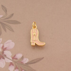 May include: A gold-toned metal charm shaped like a cowboy boot, featuring a light pink enamel inlay and a small star detail on the shaft. The charm includes a small circular loop at the top for attachment to a necklace or bracelet chain.
