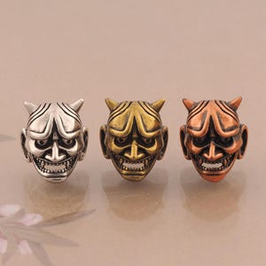 May include: Three metal beads shaped like Hannya masks, each featuring a distinct finish. One bead is silver-toned, one is gold-toned, and one is copper-toned. Each mask displays detailed facial features, including horns, wide eyes, and bared teeth.