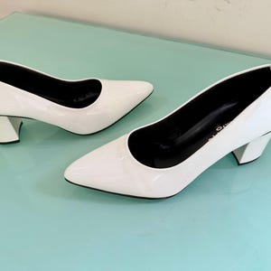 May include: A pair of white patent leather pumps with a pointed toe and a block heel. The interior of the shoes is black, and the exterior features a glossy finish. The shoes have a low-cut vamp and a structured silhouette with a solid white heel.