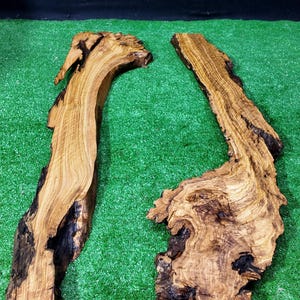 Op de afbeelding: Twee onregelmatig gevormde houten planken met een levendige rand en een natuurlijke, organische uitstraling. Het hout heeft een warme, bruine tint met donkere, bijna zwarte accenten. De planken worden op een groene ondergrond gepresenteerd.