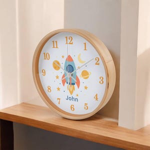 May include: A round wooden clock with a white face featuring a rocket ship, planets, stars, and the name "John". The clock has orange numerals and hands, and a light brown wooden frame.