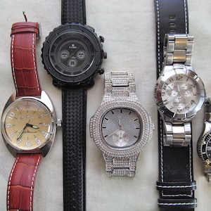 May include: Five wristwatches with different styles and designs. The watches have black, brown, and silver bands. The watch faces are round and have different colored backgrounds, including black, white, and cream.