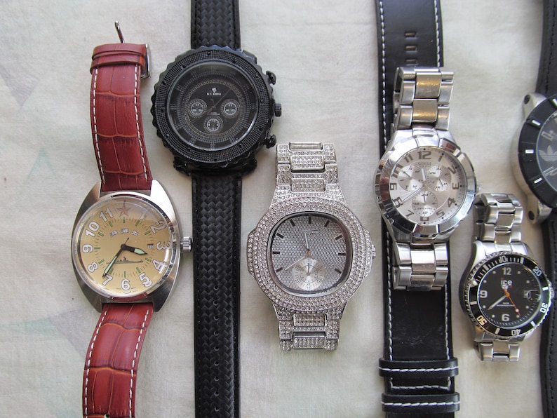 May include: Five wristwatches with different styles and designs. The watches have black, brown, and silver bands. The watch faces are round and have different colored backgrounds, including black, white, and cream.