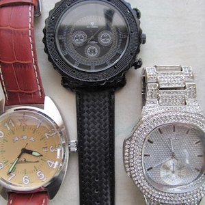 May include: Three wristwatches with different styles and designs. The first watch has a brown leather band and a round face with a beige background. The second watch has a black band with a textured pattern and a round face with a black background. The third watch has a silver band with a diamond-like pattern and a round face with a white background.