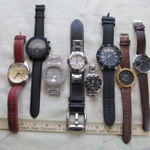 May include: A collection of eight wristwatches with different styles and designs. The watches have various colored bands, including black, brown, and red leather, and a silver metal band. Some of the watches have round faces, while others have square or rectangular faces.