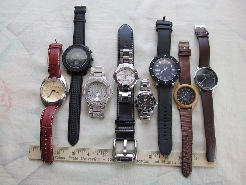 May include: A collection of eight wristwatches with different styles and designs. The watches have various colored bands, including black, brown, and red leather, and a silver metal band. Some of the watches have round faces, while others have square or rectangular faces.