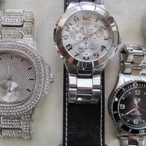 May include: Four wristwatches with silver and black bands. The watch on the left has a silver face with a diamond-like design. The watch in the center has a silver face with black numbers and hands. The watch on the right has a black face with silver numbers and hands. The watch in the background has a black face with silver hands.