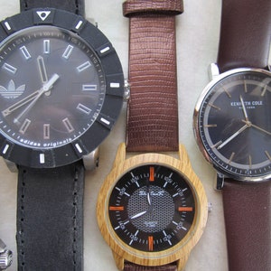 May include: Four wristwatches with different styles and designs. The watches have black, brown, and wood-toned bands. One watch has a black face with white markings and the Adidas logo. Another watch has a black face with gold markings and the text "Kenneth Cole New York". The third watch has a black face with orange markings and a wood-toned band. The fourth watch has a black face with silver markings and a silver band.