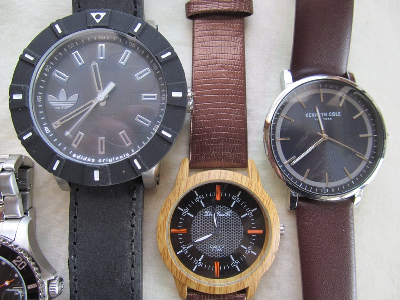 May include: Four wristwatches with different styles and designs. The watches have black, brown, and wood-toned bands. One watch has a black face with white markings and the Adidas logo. Another watch has a black face with gold markings and the text "Kenneth Cole New York". The third watch has a black face with orange markings and a wood-toned band. The fourth watch has a black face with silver markings and a silver band.