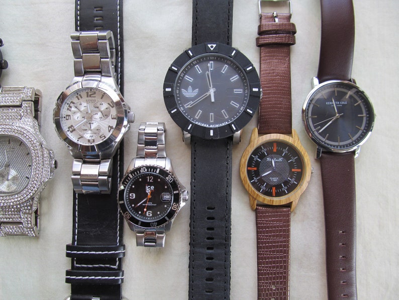 May include: A collection of six wristwatches with different styles and designs. The watches feature various band materials, including leather, metal, and wood. Some of the watches have a silver or gold tone, while others have a black or brown tone.