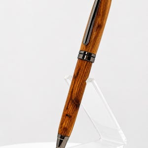 May include: A ballpoint pen featuring a polished wood barrel with a distinct brown grain pattern, accented by dark gunmetal hardware including a pocket clip, center band, and conical tip, displayed at an angle on a clear acrylic stand.