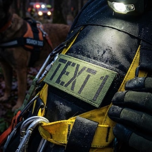 Könnte beinhalten: Nahaufnahme eines schwarz-gelben Geschirrs mit dem Text "TEXT 1" auf einem grünen Aufnäher. Das Geschirr hat silberne Karabiner und wird von einer Person in einer Waldumgebung getragen. Ein Hund mit einer "SEARCH DOG"-Weste ist im Hintergrund.