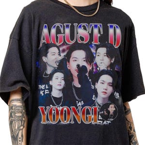 May include: Dark gray t-shirt featuring a graphic of a person with the text "AGUST D" and "YOONGI" in a retro style. The shirt has multiple images of the person in various poses. The text "THE L KS F" is also visible.
