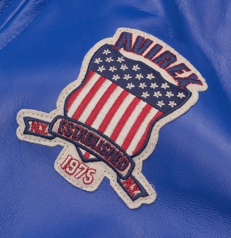 May include: Close-up of a blue leather jacket with a patch. The patch features the American flag with red and white stripes and white stars. The patch also includes the text "AVIREX ESTABLISHED 1975 NY".