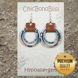 May include: A pair of dangle earrings featuring a brown leather-textured rectangular top piece connected to two concentric circular rings with a blue, orange, and white patterned finish. The earrings are displayed on a card labeled ChicBohoBliss.