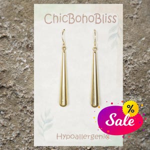 May include: A pair of gold-toned drop earrings featuring long, smooth, tapered cylindrical pendants suspended from thin wire hooks. The earrings are displayed on a white card labeled ChicBohoBliss and Hypoallergenic with a colorful Sale graphic.