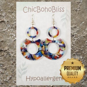 May include: A pair of dangle earrings featuring a multi-colored confetti pattern in shades of blue, red, yellow, and white. Each earring consists of a small circle connected to a larger crescent-shaped ring, suspended from gold-tone hooks.