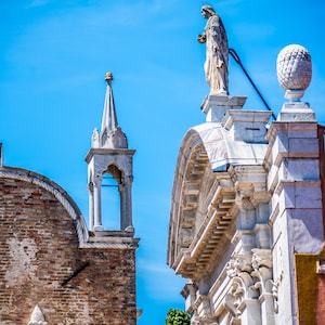 May include: Architectural details of a building against a bright blue sky. The image features a statue atop a structure, a decorative finial, and a brick facade. The building's ornate design and the clear sky create a striking contrast.