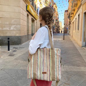 May include: A large tote bag with vertical stripes in various colors, including red, yellow, and blue, is carried over the shoulder. The bag has tan-colored straps and a small brown label. The background shows a European street scene with buildings and colorful flags.