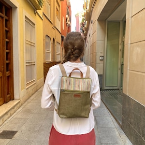 Può includere: Uno zaino verde oliva con un pannello superiore a righe e una piccola etichetta in pelle marrone. Lo zaino ha spallacci regolabili e una maniglia superiore. La persona indossa una camicia bianca e pantaloni rossi, camminando per una strada stretta.