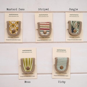 May include: Five cable holders by aseismanos on individual cards labeled Mustard fans, Striped, Jungle, Moss, and Vichy. Each holder features a fabric center with a metal snap button, framed by a beige border, designed and handcrafted in Spain.
