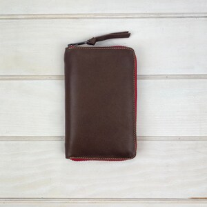 May include: A rectangular dark brown leather wallet featuring a full-length zipper closure with a matching leather pull tab. The zipper tape is a vibrant red color, providing a distinct contrast against the smooth, dark brown leather exterior.