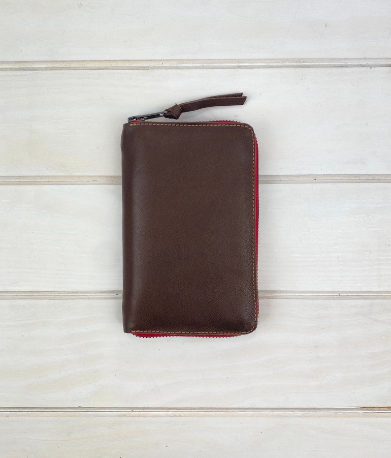 May include: A rectangular dark brown leather wallet featuring a full-length zipper closure with a matching leather pull tab. The zipper tape is a vibrant red color, providing a distinct contrast against the smooth, dark brown leather exterior.