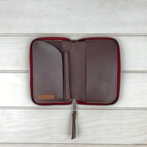 May include: A dark brown leather zip-around folio with a red interior lining. The interior features a curved pocket on the left, a vertical pen loop in the center, and a full-length pocket on the right. A leather pull tab is attached to the zipper.