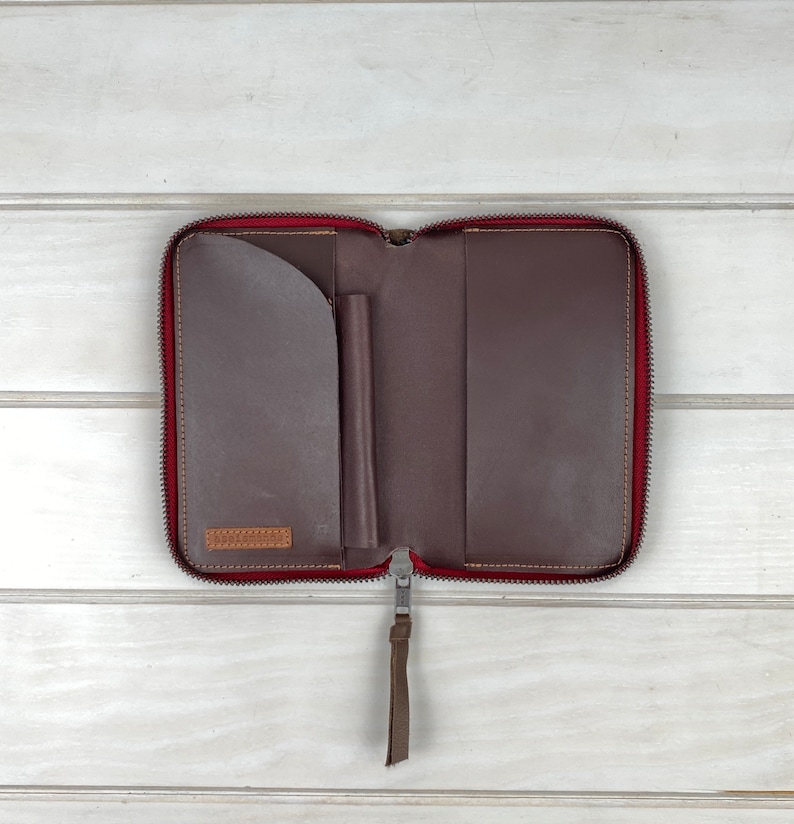 May include: A dark brown leather zip-around folio with a red interior lining. The interior features a curved pocket on the left, a vertical pen loop in the center, and a full-length pocket on the right. A leather pull tab is attached to the zipper.