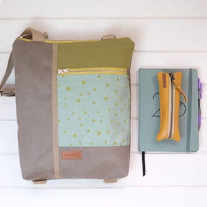 Puede incluir: Un bolso bandolera vertical con un panel lateral gris, una sección superior verde y un bolsillo frontal azul claro con lunares amarillos. Al lado hay un cuaderno azul claro y un estuche amarillo mostaza con cremallera y tirador de cuero.