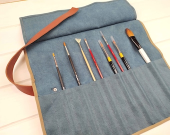May include: A blue canvas roll-up case featuring a brown leather strap and multiple interior slots. Seven paint brushes with varying tip shapes and handle colors, including black, red, and white, are inserted into the individual fabric pockets.