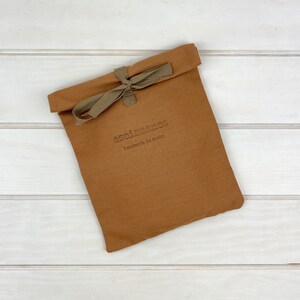 May include: A rectangular, tobacco-brown fabric pouch with a folded top edge secured by a tied olive-green ribbon. The front of the pouch features the embossed text "aseismanos" followed by the smaller text "handmade in spain" in a dark font.