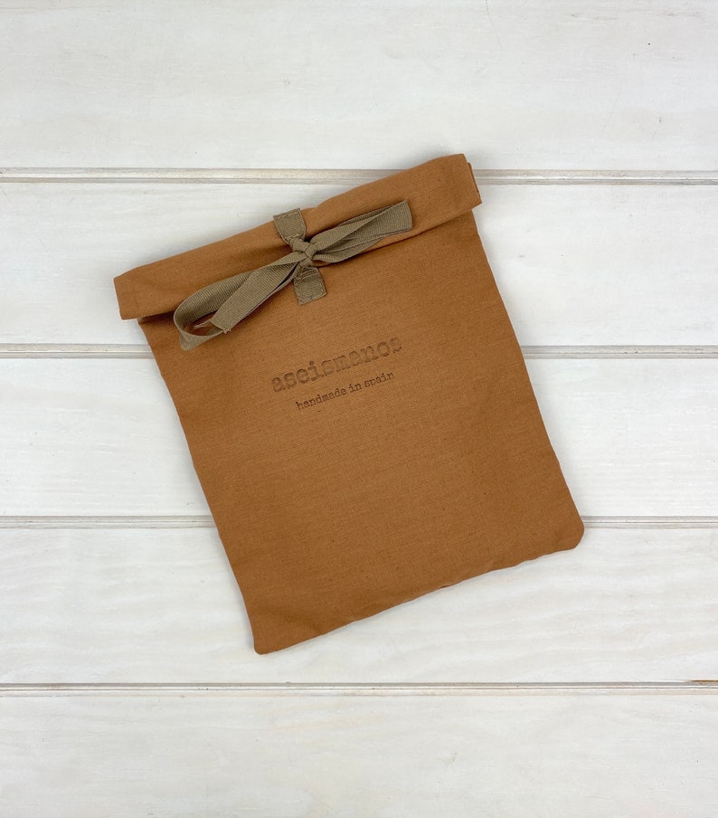 May include: A rectangular, tobacco-brown fabric pouch with a folded top edge secured by a tied olive-green ribbon. The front of the pouch features the embossed text "aseismanos" followed by the smaller text "handmade in spain" in a dark font.