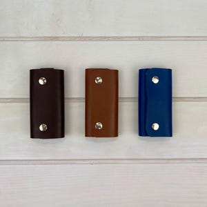 May include: Three rectangular key cases in dark brown, tan, and blue. Each case features a smooth finish and is secured by two silver-toned metal snap buttons positioned vertically along the center of the front flap for closure.