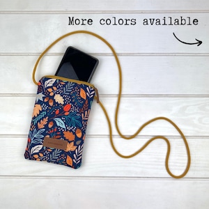 Puede incluir: Una pequeña bolsa azul para teléfono con un estampado de hojas de otoño coloridas. La bolsa tiene una correa larga amarilla y un teléfono negro en el interior. "Más colores disponibles" está escrito encima de la bolsa.