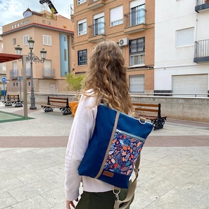 Könnte beinhalten: Ein blauer, wandelbarer Rucksack in rechteckiger Form mit einer vorderen Reißverschlusstasche, die ein buntes Herbstblattmuster aufweist. Die Tasche verfügt über verstellbare beige Riemen, silberne Metallbeschläge und ein kleines braunes Etikett.